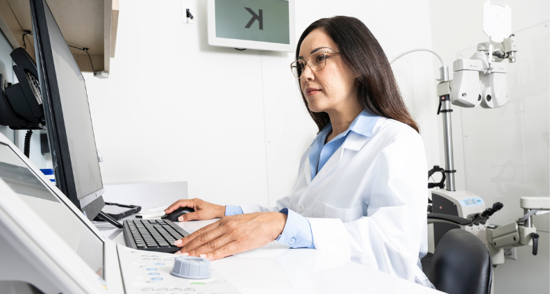 A female optometrist is sitting in an exam room working on a computer.