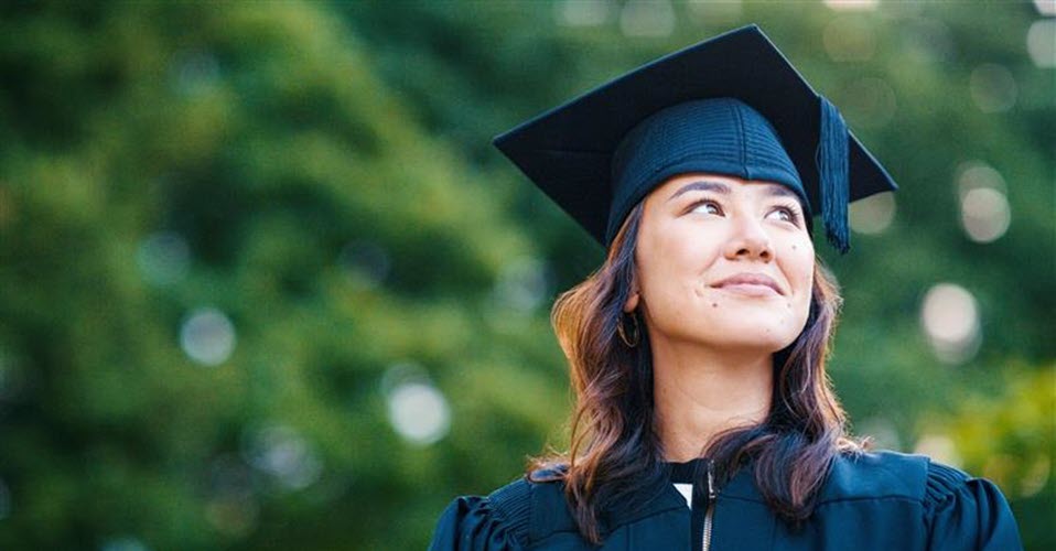 A woman with brunette hair wears a dark green graduation cap and gown