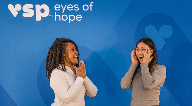 Two women standing in front of a blue heart-patterned background with the VSP Eyes of Hope logo with one woman watching the other excitedly putting on a new pair of glasses