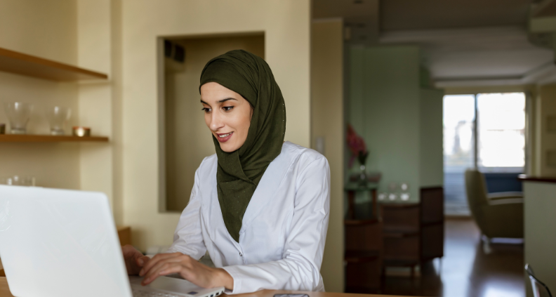 A woman, wearing an olive-colored hijab and a white blazer, is sitting at a table and typing on a white laptop.