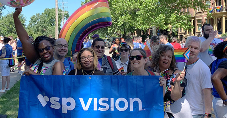 VSP Vision employees are holding a blue banner with "VSP Vision" in white text while walking in the City of Columbus, Ohio, Pride festival. A rainbow balloon is in the background.