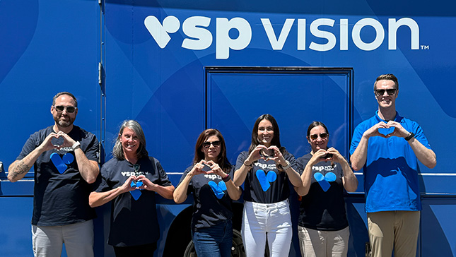 Six VSP Vision employees volunteering at an Eyes of Hope vision care outreach event are standing from left to right in front of a VSP Eyes of Hope mobile clinic, holding their hands up in the shape of a heart. They're wearing blue shirts with VSP Vision branding.