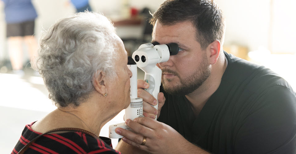 A volunteer optometrist delivers a comprehensive eye exam to a patient at an Eyes of Hope mobile clinic event in Lafayette, Louisiana. The patient is an adult with short gray, curly hair and the optometrist has short dark hair with a mustache and beard and is wearing black scrubs.