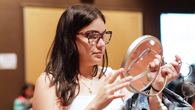 VSP Eyes of Hope patient tries on new eyewear at a free vision care event with Visionworks in San Antonio, Texas. The patient has long brown hair and is wearing a white shirt and tortoiseshell frames while looking in a small round handheld mirror with another pair of frames in her hand.
